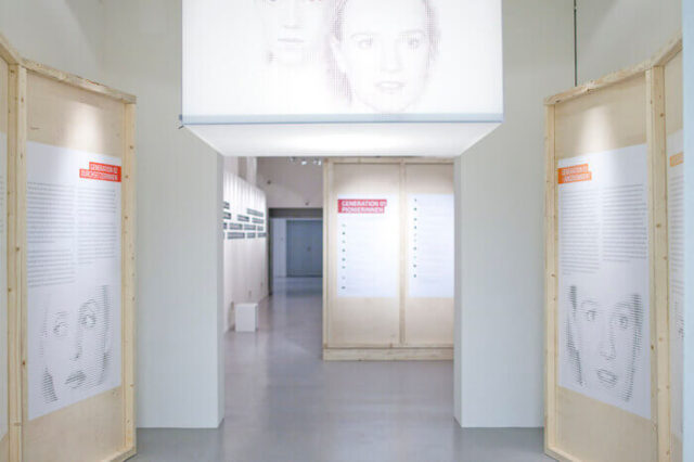 Ausstellung mit Holzstelen und projizierten Porträts in einem hellen Gang